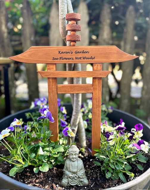 Wooden garden sign with engraved text in a pot with flowers and a small statue.
