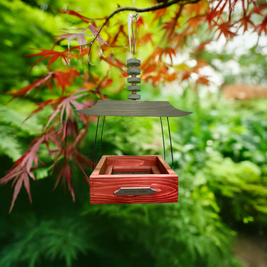 Wooden Asian style bird feeder hanging from a branch with a blurred green and red foliage background