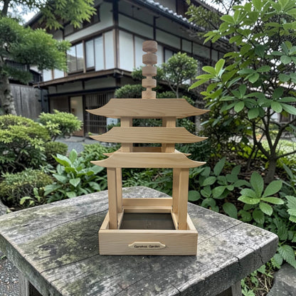 Wooden pagoda-shaped bird feeder on a stone surface with greenery and traditional building in the background