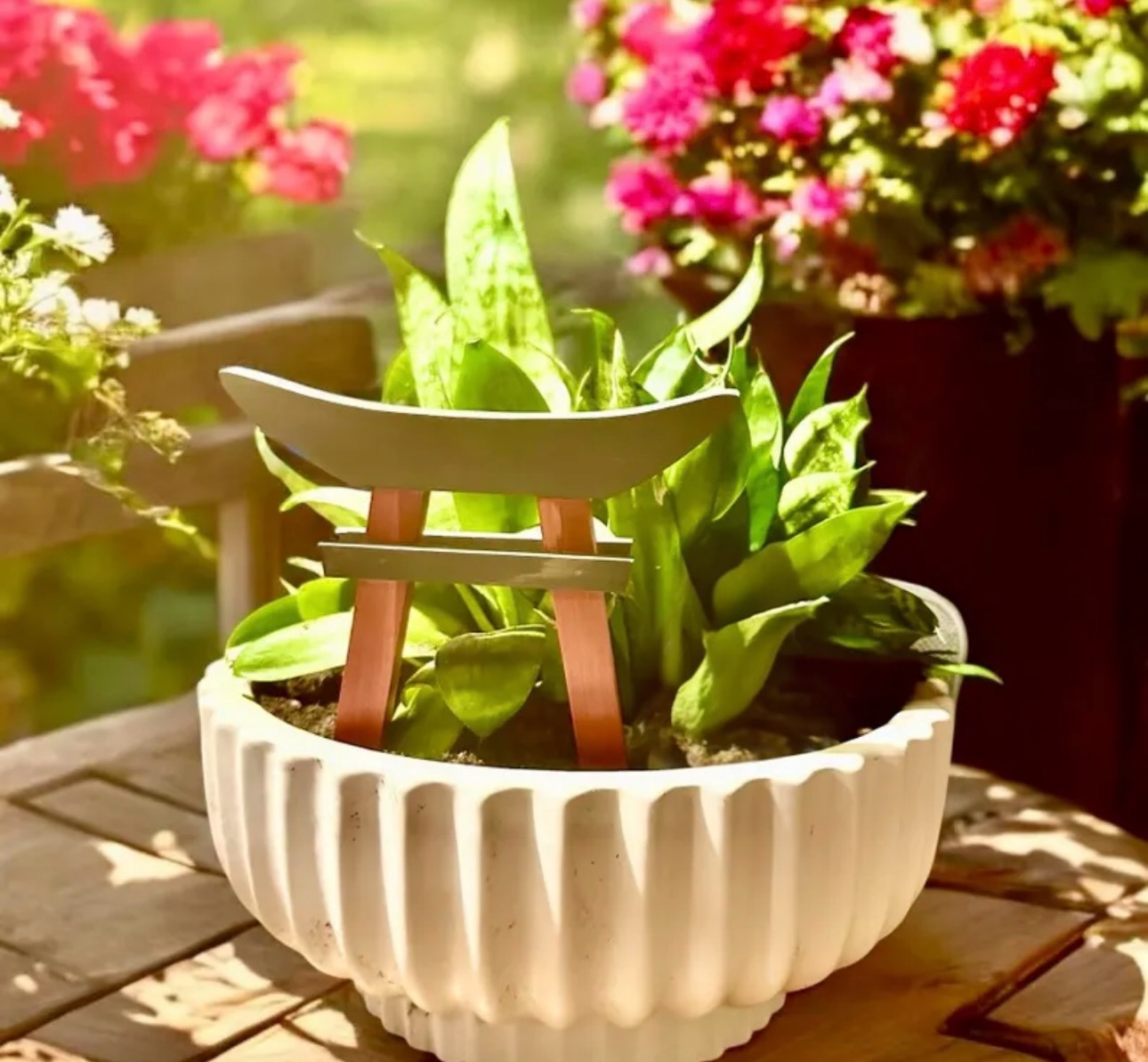 White decorative planter with red and grey mini Torii Gate planter stake on a wooden surface with blurred flowers in the background.